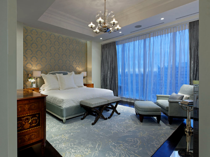This bedroom design by Douglas Design Studio is the perfect combo for antique pieces such as the side tables and sideboard in wood with a more modern bed and chaise longue in blue combined with a rug in the same blue that covers the room.