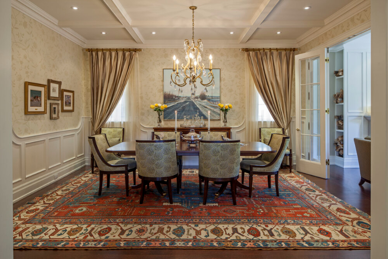 This dining room designed by Douglas Design Studio is a space that evokes antique charm such as the chandelier and the armchairs in beige palette. The room is covered by a rug in red and beige that contrasts with the white and beige of the walls.