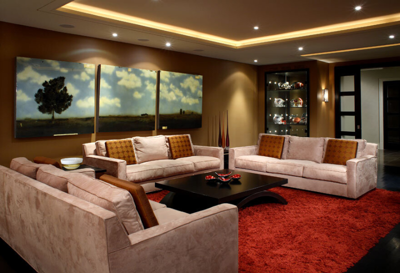 This dark living room is designed by Douglas Design Studio and includes three sofas in beige that contrasts with the enormous red rug that covers all the tree sofas. The brown walls show tree paintings of a field with a tree.