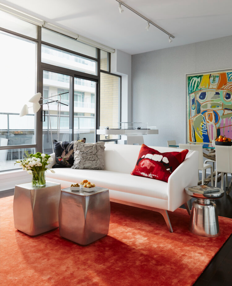 This living room is designed by Douglas Design Studio and as the centerpiece of the room is a rug in orange/red color that combines with a white sofa and the two coffee tables in the center of the room in "metal".