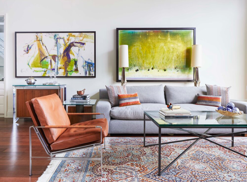 This converted boutique loft was designed by Douglas Design Studio and has a grey sofa with orange/brown pillows that are combined with the armchair in the same color as the pillow. The room shows two paintings in green and yellow colors. The room is covered with a rug in blue, orange, red, and white colors.