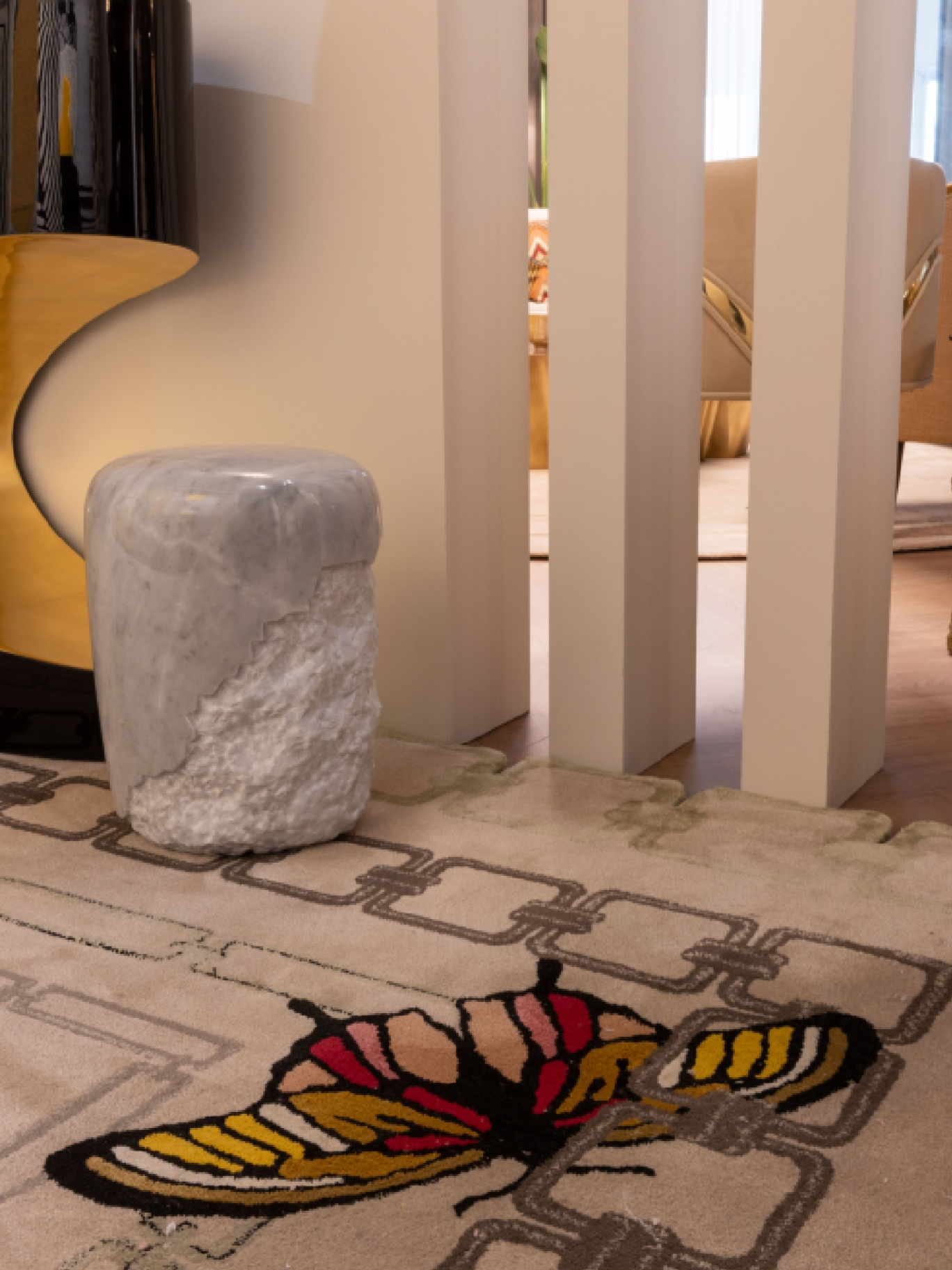 A luxury interior design detail featuring a sculptural white marble stool with a rough-hewn base on a cream-colored rug with a large, colorful butterfly illustration and chain-link pattern, set beside beige architectural pillars and a gold metallic furniture piece.
Rug'Society at Salone del mobile 2026