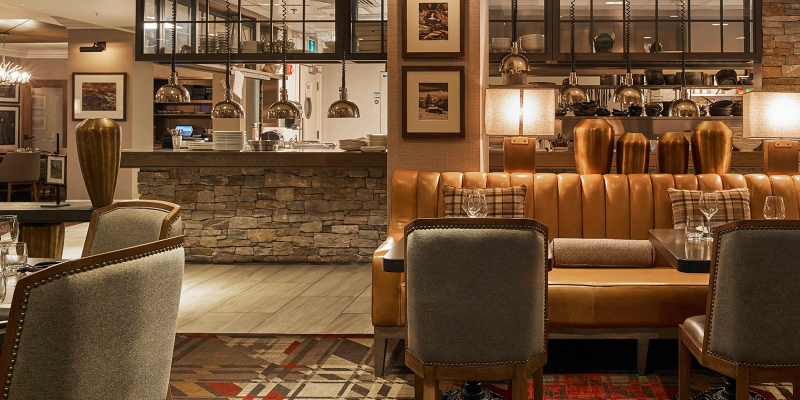 Rug ideas with Navigate Design. This restaurant has a brown leather sofa, wood tables, grey dining armchairs, and a rug in red, brown, and white.