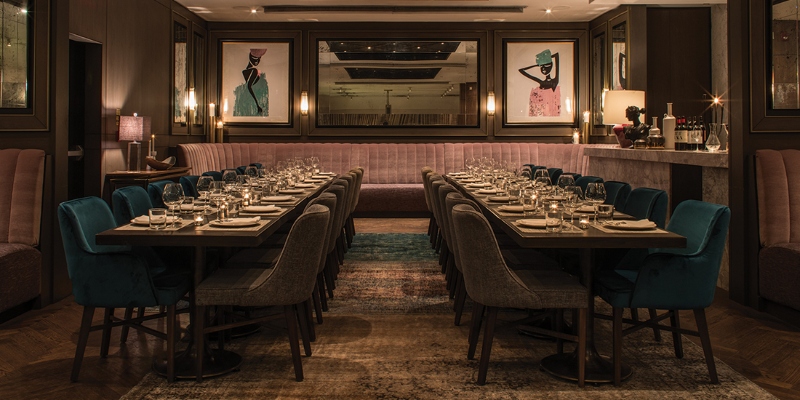 Rug ideas with Navigate Design. This restaurant with a dark ambiance has a big round red sofa on the opposite wall and there are two big tables, dining armchairs in blue and grey, and a rug with dark colors.