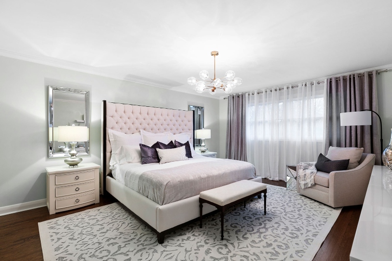 Rug Design Inspiration by Camden Lane Interiors. This bedroom with grey walls has a white rug, a light pink armchair and bed frame, two side tables in white and at the end of the bed is a bench in white.