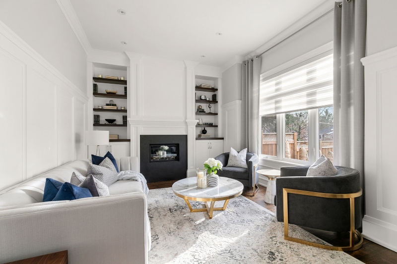 Rug Design Inspiration by Camden Lane Interiors. This white living room with shelves in wood and a black fireplace has a white sofa, two dark grey armchairs, a coffee table with marble countertop and gold structure and covering all the room has a white rug with light grey colors.