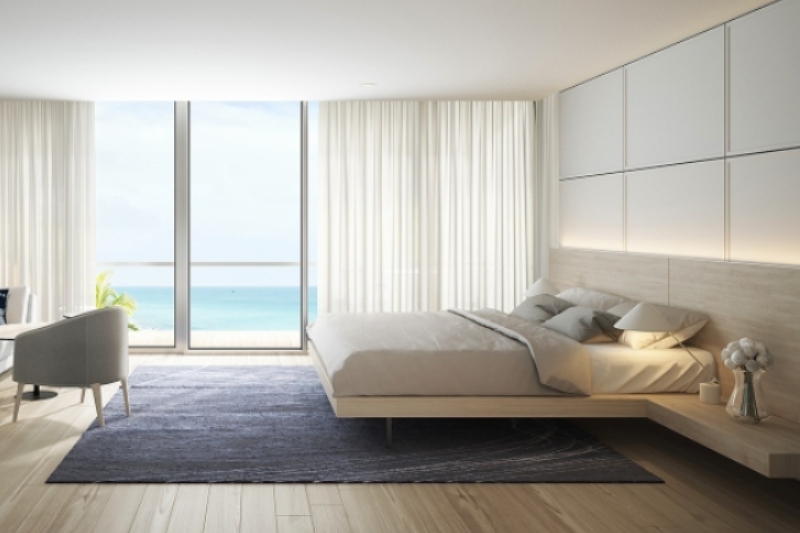 This white modern bedroom has a bed frame in light wood. a blue rug, a white lounge, and one grey armchair.